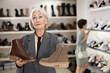 © JackF - Senior female customer is standing next to shelf in stores sales area. She picks up shoes, looks at and studies material and quality of product, decides to buy shoes