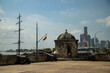 © Leandro Lima - Contraste entre o novo e o antigo, com prédios altos e modernos ao fundo e uma torre de uma fortaleza com uma fragata atracada no mar caribenho.