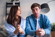 © simona - Young couple sitting together on a sofa at home, engaged in a tense conversation. Expressions show frustration, concern and distance as they discuss relationship issues and try to communicate.