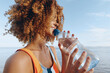 © SHOTPRIME STUDIO - A smiling woman with curly hair drinks water from a plastic bottle outdoors near the sea. The woman enjoys a healthy lifestyle with fresh air and natural light.