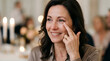© Alena - Woman with dark hair wearing a beige blouse touches her face while smiling at a luxury wedding reception with soft candlelight and blurred guests in the background