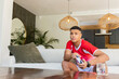 © wavebreak3 - Man sitting in open-plan living room wearing red sports shirt holding game controller, copy space