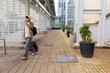 © wavebreak3 - Asian man walking covered corridor pulling black rolling suitcase carrying backpack drinking cup