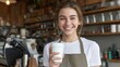 © Iuliia - Smiling woman barista holding white paper coffee cup in cafe. Young caucasian employee offering drink to customer. Professional coffee shop worker providing service in modern restaurant interior.
