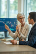 © jina choi - Confident mature senior businesswoman discussing financial charts with a younger male colleague in a modern office