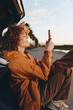 © SHOTPRIME STUDIO - Smiling woman with curly hair wearing an orange jacket sitting by car trunk using smartphone outdoors during sunset. Lifestyle concept with casual outfit and natural light mood.