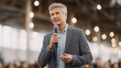 © Ally_art - Gray-haired man speaker giving a speech with a microphone at a business conference. Professional keynote orator talking to audience in a convention hall. Leadership and corporate event.