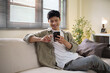 © matinee - Young Asian man relaxing on a sofa while using a smartphone at home. Concept of online communication, mobile technology, and modern lifestyle.