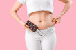 © Pixel-Shot - Young woman in tight pants with chocolate on pink background, closeup. Weight gain concept