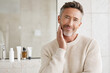 © kora studio - Healthy looking older man in his fifties with neatly styled hair smiles while touching his face in modern bathroom setting. conveys confidence and self care