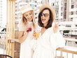 © halayalex - Two young beautiful smiling hipster female in trendy summer white dress and straw hats. Sexy carefree women holding and drinking fresh vegetable cocktail smoothie drink in plastic cup with straw