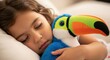 © Olga Ко - Menina adorável dormindo pacificamente abraçada a um tucano de pelúcia colorido, conceito de sono infantil e conforto