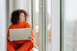 © Westend61 - Smiling woman with curly hair using laptop on windowsill indoors