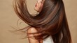 © Newleks - Young woman confidently tossing her long, lustrous brown hair against a neutral background, creating a dynamic visual of movement, healthy locks, and glamorous beauty