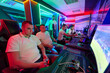 © .shock - Young Men Gaming Together in Neon-Lit LAN Center Playing Console and PC Multiplayer Games