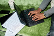 © Mediaphotos - High angle view of Black young professional typing on laptop beside documents and smartphone on grass, working outdoors. Useful for remote work, productivity, freelance business marketing