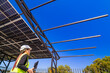 © ultramansk - A female engineer in a safety vest and helmet inspecting a solar carport under construction. The structure’s steel beams and partially installed solar panels are visible under a bright blue sky.