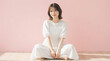 © k-studio - Young Japanese woman sitting cross-legged in a white dress on pink background