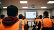 © Sulfabndi - Group of people attending a training session or workshop in a modern classroom setting with a large screen at the front and participants wearing safety vests