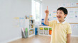 © Alena Yakusheva - Cheerful boy pointing up while standing in a bright modern classroom, active participation, inclusive education, primary school student portrait.