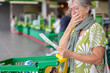 © luciano - Worried senior woman standing in a supermarket checkout area, holding and reviewing her shopping receipt beside a grocery cart, expressing concern about household expenses and rising food prices
