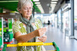 © luciano - Senior woman standing in a supermarket checkout area, holding and reviewing her shopping receipt beside a grocery cart, expressing concern about household expenses and rising food prices