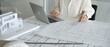 © volha_r - Architect working on new residential project designs, using laptop and reviewing blueprints with an architectural model and safety helmet on planning table