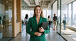 © Vyatcheslav - Professional woman in a green blazer holding a folder in a modern office. Smiling businesswoman in a corporate hallway. Career and leadership concept