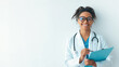 © Prostock-studio - A young female doctor stands by a white wall in an office. She smiles while holding a clipboard. She wears a white coat and has glasses on. Her stethoscope hangs around her neck.