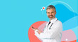 © Prostock-studio - A middle aged doctor stands smiling against a blue background. He points to open space, suggesting information about checkup plans at his clinic. He looks approachable and friendly.