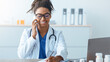 © Prostock-studio - A doctor is on a phone call while seated at a desk. She is wearing a white coat and glasses. Medical supplies are visible on the desk. A laptop is present in the background.