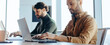 © Prostock-studio - Two men sit at a long desk working on their laptops. They focus on their tasks in an office filled with natural light. A coffee cup and a phone are on the table.