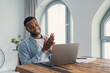 © Daniel - A smiling man applauds in front of his laptop, celebrating a successful remote work moment in a bright, modern home office. Papers and a notebook sit on the wooden table beside him.