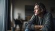 © Kelapa - A contemplative middle-aged man gazes out a window, his expression suggesting introspection and a moment of quiet solitude within a modern, dimly lit interior.