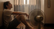 © NeuroCake - Young man suffering from a summer heatwave at home. Exhausted person sweating and cooling down with an electric fan during hot weather