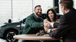 © wedmoments.stock - Couple discussing auto deal with salesman, shaking hands at a table inside a modern car dealership, vehicle visible in the background