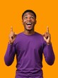 © Vasiliy - Vertical portrait of an excited young Black man pointing up. Happy African American male in a purple shirt looking upwards in surprise against an orange background. Copy space for text
