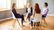 © Rahman - Blurry image of a group of people sitting in chairs in a circle talking to each other in a room with gray walls and wooden floors
