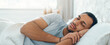 © Prostock-studio - A man lies in bed with his eyes closed, resting on a pillow. Sunlight enters the room, creating a warm atmosphere. He appears comfortable and relaxed while sleeping in a cozy setting.