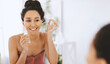 © Prostock-studio - A woman smiles while holding a skincare product in front of a mirror. She is in a bright room, focusing on her skincare routine. Sunlight enters through the window.