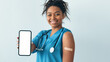 © Prostock-studio - A health care worker in a blue uniform smiles while holding a smartphone. She has a bandage on her upper arm after receiving a vaccination. The setting is a health clinic.