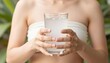 © Tati - Young woman holding a glass of iced water with both hands, torso visible in a white strapless top against a softly blurred green foliage background