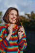 © SHOTPRIME STUDIO - Woman smiling in a colorful striped sweater holding sunglasses outdoors; young woman portrait with bob haircut, casual outfit and joyful mood, lifestyle fashion shot in warm sunlight.