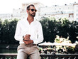 © halayalex - Handsome confident stylish hipster lambersexual model.Sexy modern man dressed in white shirt and trousers. Fashion male posing in the street near embankment in Europe park city at sunset.In sunglasses