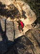 © VICTOR - Rock climber in bright red shirt ascends rugged stone face. Sunlight creates sharp shadows across textured rock surface. Red climbing rope secures hiker's position firmly