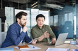 © Liubomir - Two diverse business professionals collaborating during a discussion, reviewing information on a laptop and tablet, working together on a project in a bright contemporary office