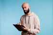 © DC Studio - Young arab man writing in clipboard with concentrated facial expression. Focused arabian bald bearded person making list and taking notes while standing on blue background