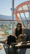 © Krakenimages.com - Woman holding smartphone and smiling while seated on a cruise terrace near glass railing inside a building with sea and mountain view; relaxation leisure.