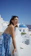 © Krakenimages.com - Woman leans on building railing at santorini church, smiling and wearing blue floral dress with sunglasses on head; vacation joy.