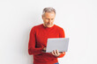 © Prostock-studio - A man stands against a white wall while he uses a laptop. He is wearing a red sweater and appears focused on his work. The setting is simple with no distractions.